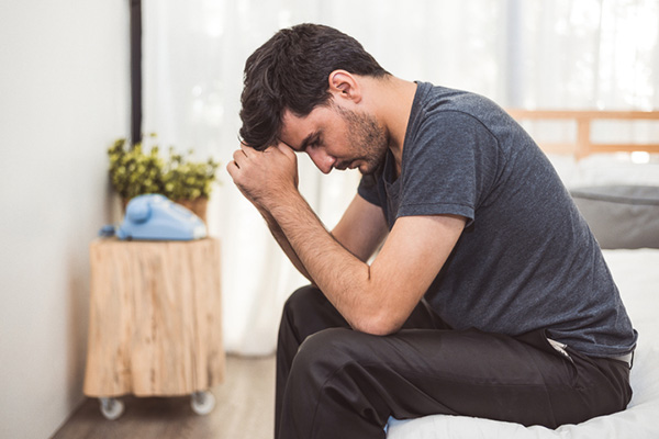 Worried man sitting on bed with hand on forehead in bedroom in serious mood emotion